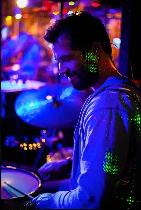Male drummer for pop rock band in France smiling to himself happily and performing at black light themed event | Altus Entertainment