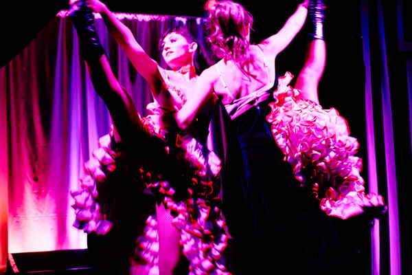 Two cabaret dancers in French cabaret show lifting one leg in air while pink lighting shines down on them | Altus Entertainment