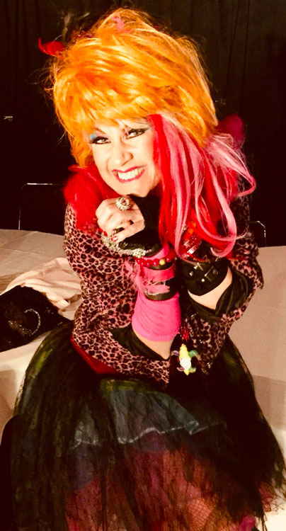 Cyndi Lauper impersonator sitting on white couch while smiling widely to camera prior to performance | Altus Entertainment