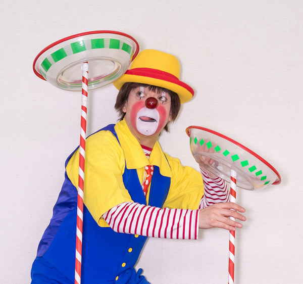 Male clown performer in Japan wearing vibrant costuming while balancing glass dishes on poles and looking to side | Altus Entertainment